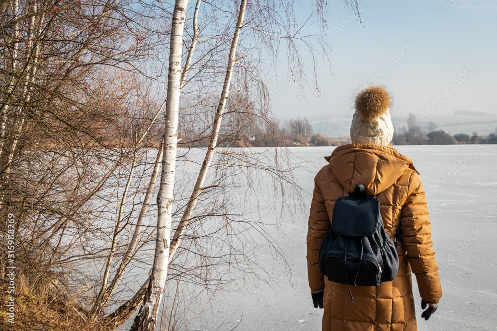 Woman wearing winter coat and knitted hat is standing at frozen lake and birch tree Woman wearing winter coat and knitted hat is standing at frozen lake and birch tree