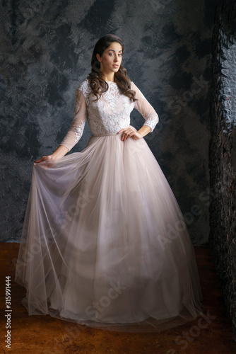 Beautiful young woman stands in a delicate wedding dress against a dark wall