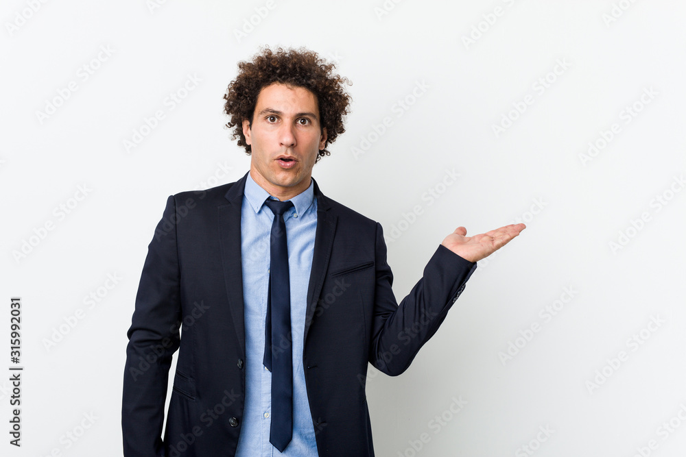 Young business curly man against white background impressed holding copy space on palm. Young business curly man against white background impressed holding copy space on palm.