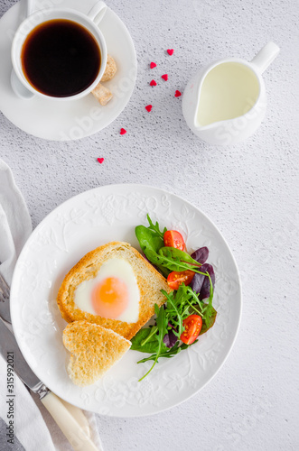Piece of bread toast cut in shape of heart with egg on a white plate with arugula and cherry tomatoes. Romantic breakfast for Valentine's Day. Top view. Vertical orientation.