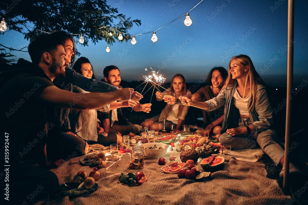 Happy friends having fun with fire sparkles. Young people millennials  camping at picnic after sunset. Stock Photo | Adobe Stock