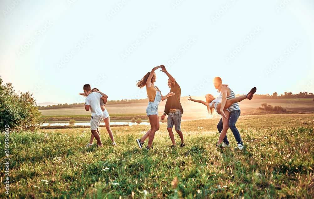 group of happy young people dancing on the field on beautiful summer sunset - obrazy, fototapety, plakaty 