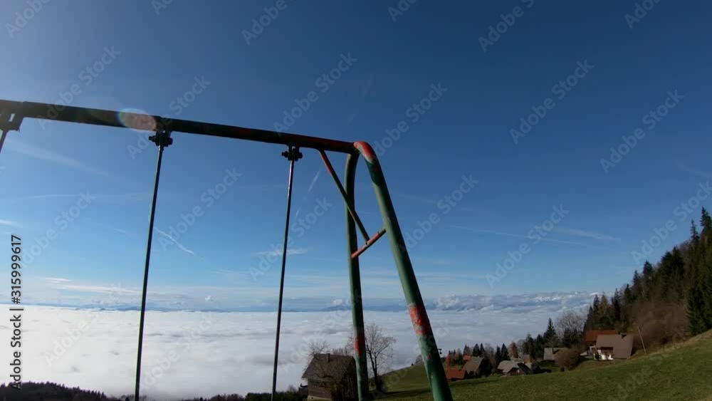 Backward moving tilt down kids swing equipment on hilltop with view on village and low altitude fog