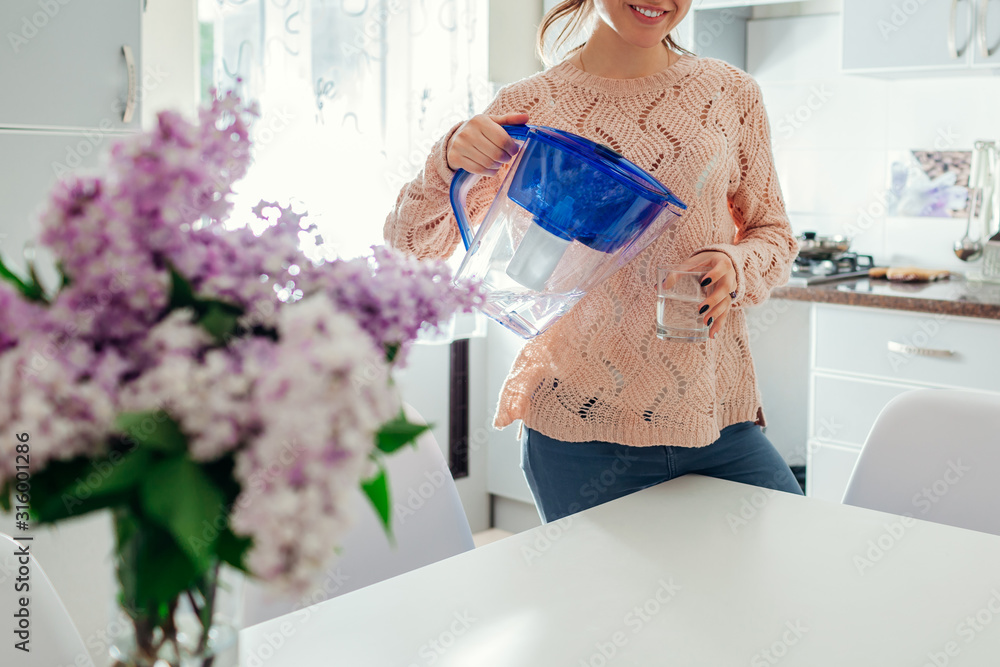Woman pouring filtered water from filter jug into glass on kitchen. Modern kitchen design. Healthy lifestyle - obrazy, fototapety, plakaty 