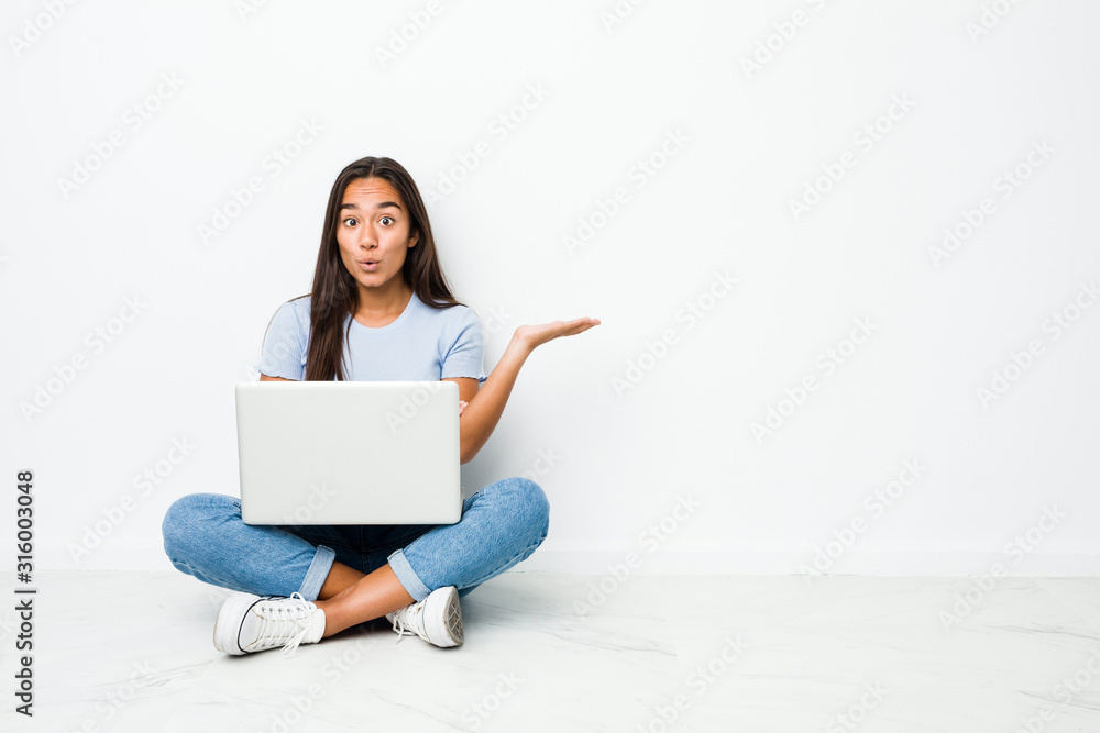 Young mixed race indian woman sitting working on laptop impressed holding copy space on palm. Young mixed race indian woman sitting working on laptop impressed holding copy space on palm.