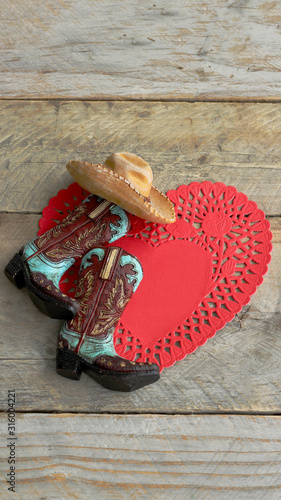 cowboy boots and hat laying on a red heart paper doily on a rustic wood background with copy space, vertical photo