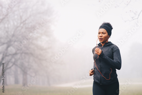 A woman merrily jogging on a misty day in nature. She is smiling. She is wearing headphones, sports clothes and a hairband.