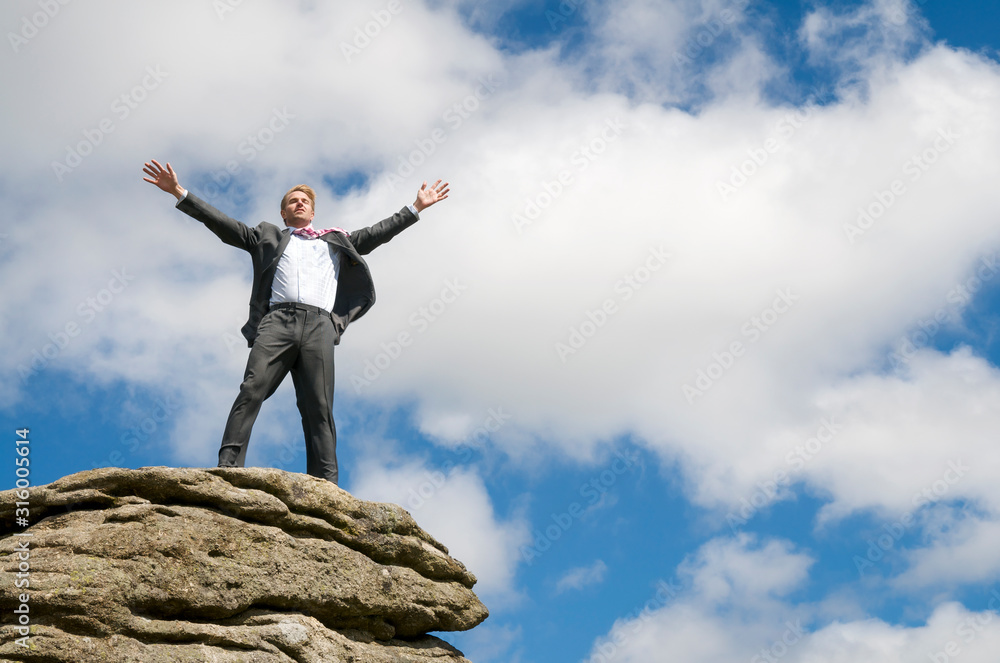 Businessman standing on the top of a distant rocky mountain peak celebrating in blue sky copy space Businessman standing on the top of a distant rocky mountain peak celebrating in blue sky copy space