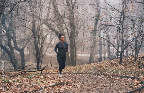 A woman merrily jogging on a misty day in nature. She is smiling. She is wearing headphones, sports clothes and a hairband.