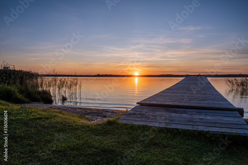 Fototapeta Naklejka Na Ścianę i Meble -  Colorful orange sunset on a quiet lake. Perspective view of a wooden pier on the pond at sunset.