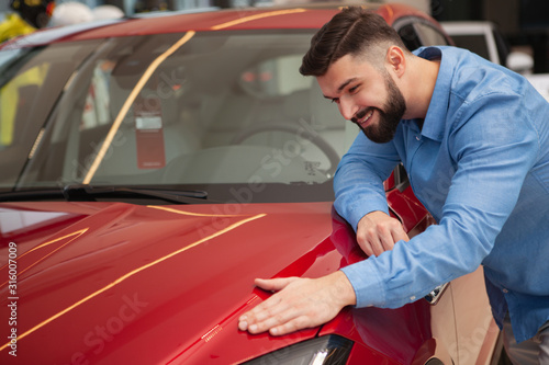 Happy handsome bearded man smiling, examining beautiful red automobile on sale at car dealership, copy space