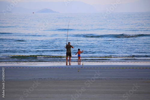 Bluish sunset at the beach. Grandfather and grandson fishing. Foggy horizon with calm waves in the sea. Itaguare beach, Bertioga, Brazil photo