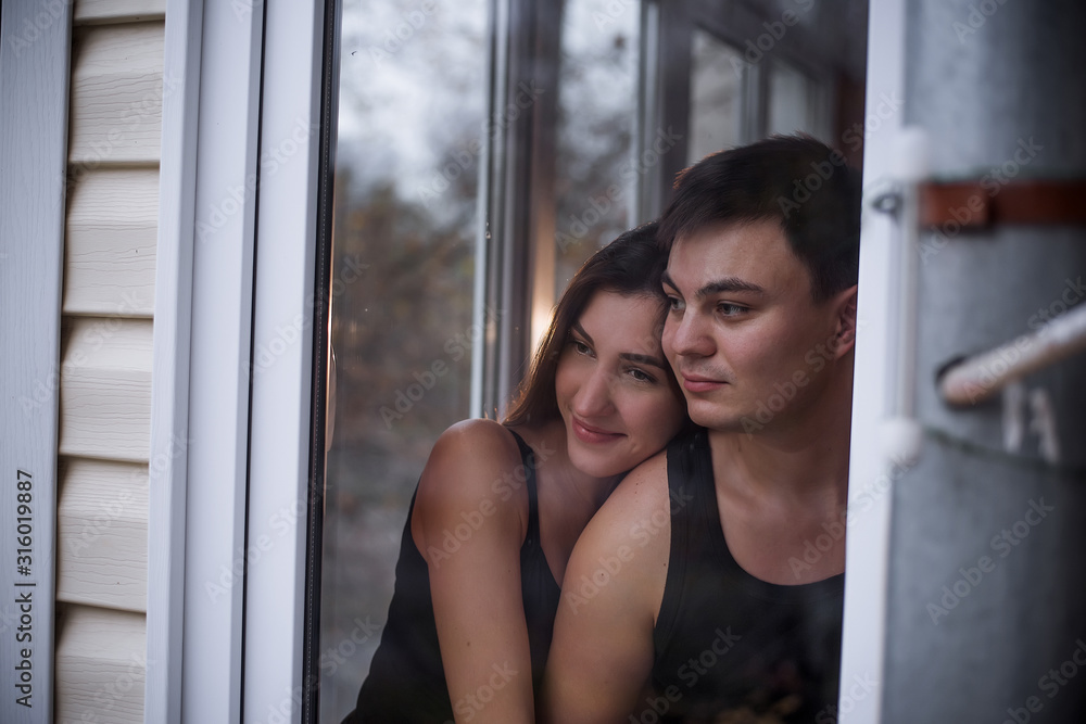 A loving young couple hugs in the window of their balcony terrace in the apartment. Young man laughs with a beautiful woman at home. Lovers kiss, passionately spend time with each other. Lifestyle