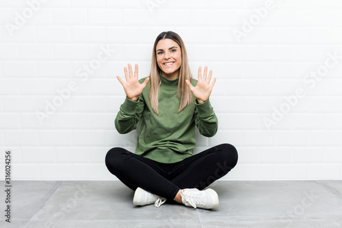 Young caucasian woman sitting on the floor showing number ten with hands.