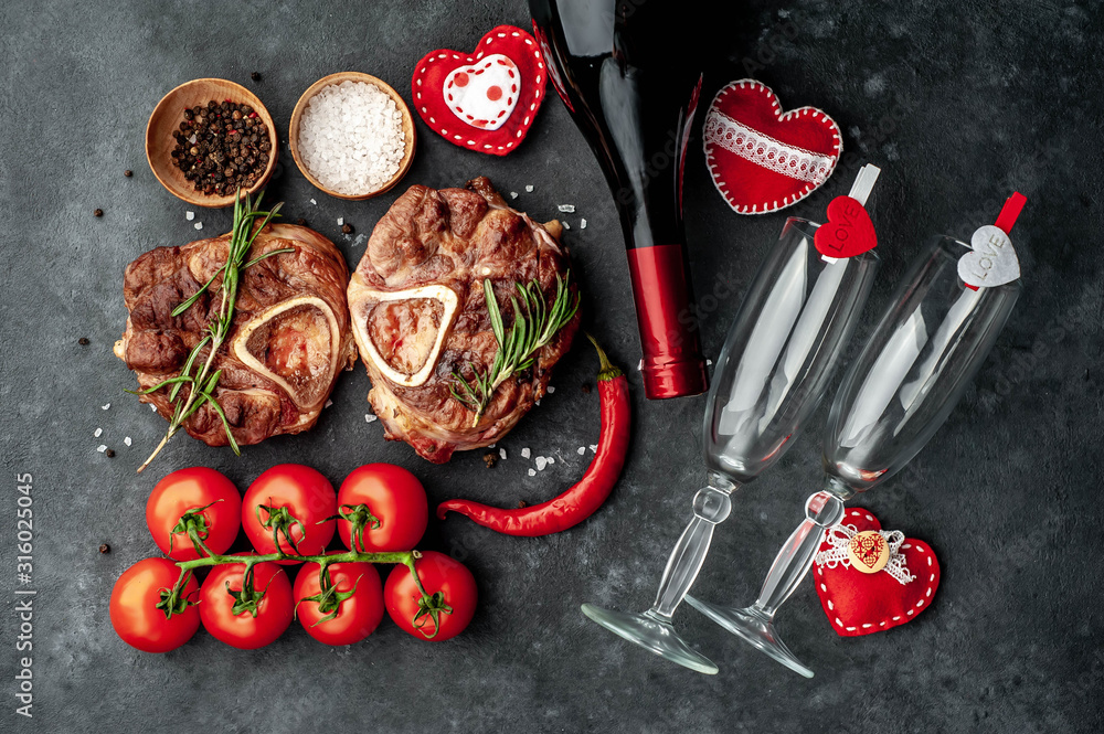 two grilled steaks, with tomatoes and spices with a bottle of wine and glasses, various red hearts on a stone background. dinner for two on valentine's day two grilled steaks, with tomatoes and spices with a bottle of wine and glasses, various red hearts on a stone background. dinner for two on valentine's day
