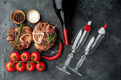 two grilled steaks, with tomatoes and spices with a bottle of wine and glasses, various red hearts on a stone background. dinner for two on valentine's day