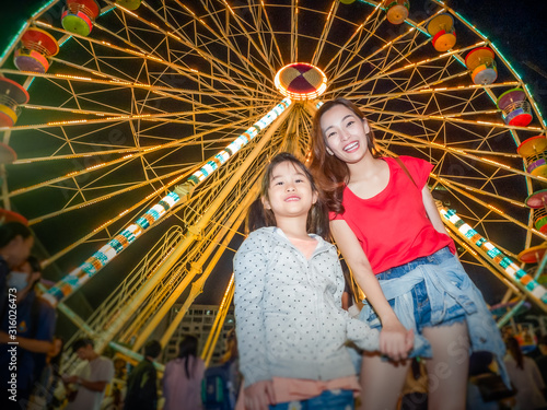 happy asia mother and daughter have fun in amusement carnival park with farris wheel and carousel background