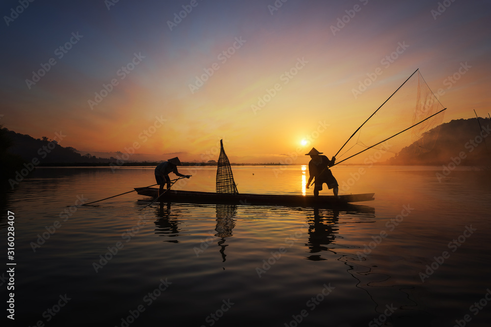 Silhouette of Asian fisherman on wooden boat in action casting a net for catching freshwater fish in nature river in the early morning before sunrise