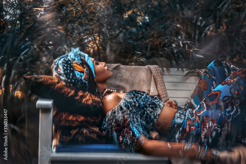 Fototapeta beautiful young stylish woman wearing turban lying on the bench in garden on tro
