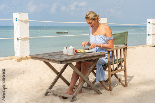 Young woman having romantic breakfast in hotel restaurant during sunrise near sea waves on the tropical beach, close up. Concept of leisure and travel
