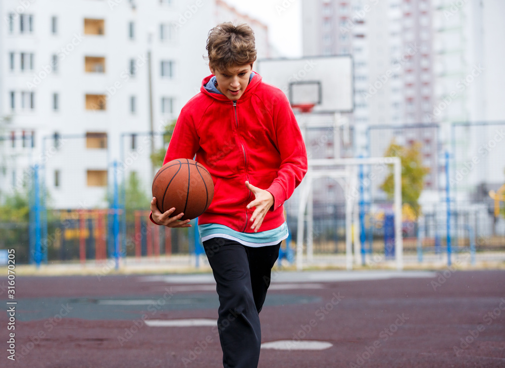 Cute boy in red t shirt plays basketball on city playground. Active teen enjoying outdoor game with orange ball. Hobby, active lifestyle, sport for kids.