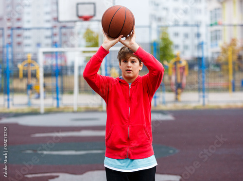 Cute boy in red t shirt plays basketball on city playground. Active teen enjoying outdoor game with orange ball. Hobby, active lifestyle, sport for kids. © Natali