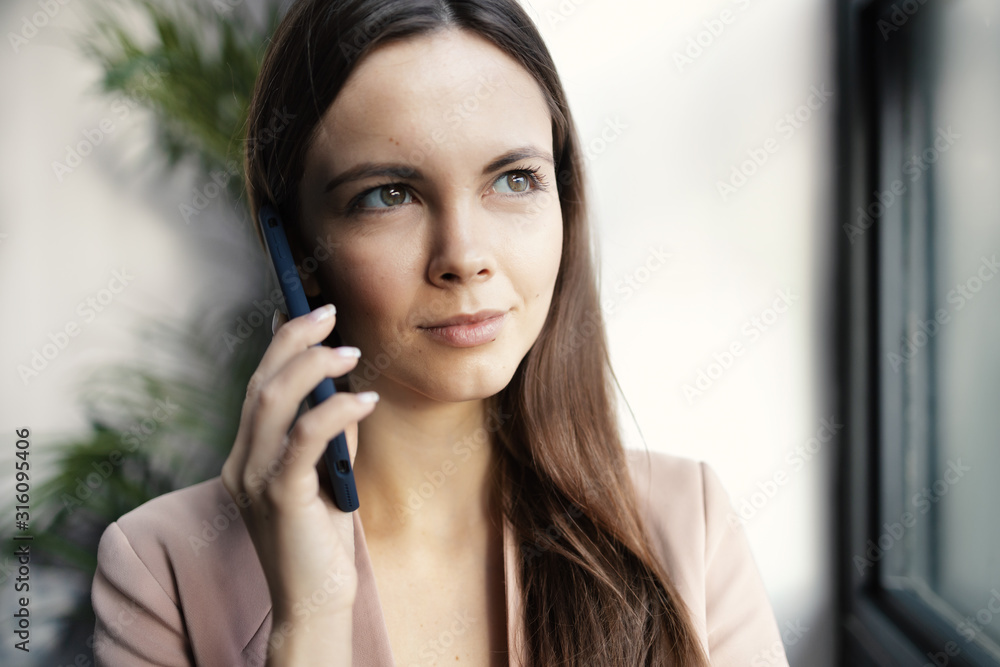 Young woman business entrepreneur in casuals standing by a window and talking over smartphone in office. Smiling attractive businesswoman talking on mobile phone with client. Young woman business entrepreneur in casuals standing by a window and talking over smartphone in office. Smiling attractive businesswoman talking on mobile phone with client.