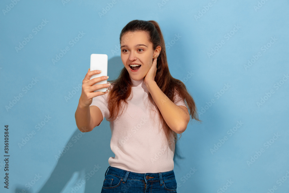 Taking selfie or vlog, shocked. Caucasian teen girl's portrait on blue background. Beautiful model in casual wear. Concept of human emotions, facial expression, sales, ad. Copyspace. Looks happy. Taking selfie or vlog, shocked. Caucasian teen girl's portrait on blue background. Beautiful model in casual wear. Concept of human emotions, facial expression, sales, ad. Copyspace. Looks happy.