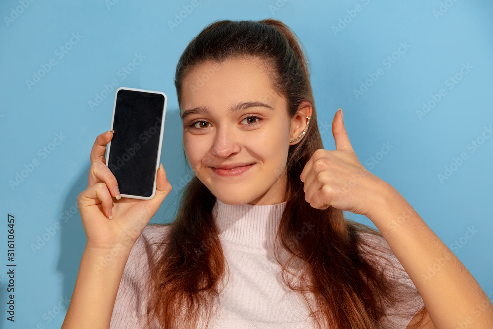 Showing phone screen, thumb up. Caucasian teen girl's portrait on blue background. Beautiful model in casual wear. Concept of human emotions, facial expression, sales, ad. Copyspace. Looks happy. Showing phone screen, thumb up. Caucasian teen girl's portrait on blue background. Beautiful model in casual wear. Concept of human emotions, facial expression, sales, ad. Copyspace. Looks happy.