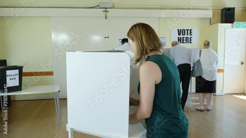 Female Voter stood at polling booth voting at Polling Place / Station. With people behind choosing who to vote for and post Ballot papers in Box photo