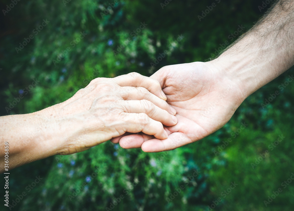 Fototapeta premium Senior and young hands outside closeup. Elderly woman and young man holding hands together, green blurred background, sunlight. Love, warmth, take care concept, Valentines, mothers day, donate, help.