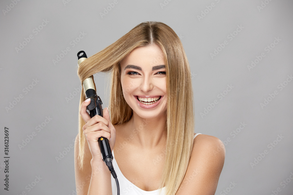 beautiful blonde woman using curling iron for her healthy long hair and smiling to camera beautiful blonde woman using curling iron for her healthy long hair and smiling to camera