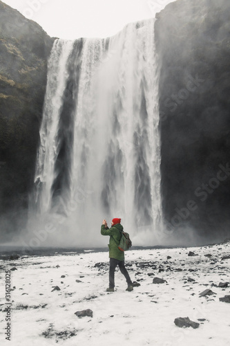 young male traveler in green clothes in a red hat and a green backpack stands near a waterfall in Iceland