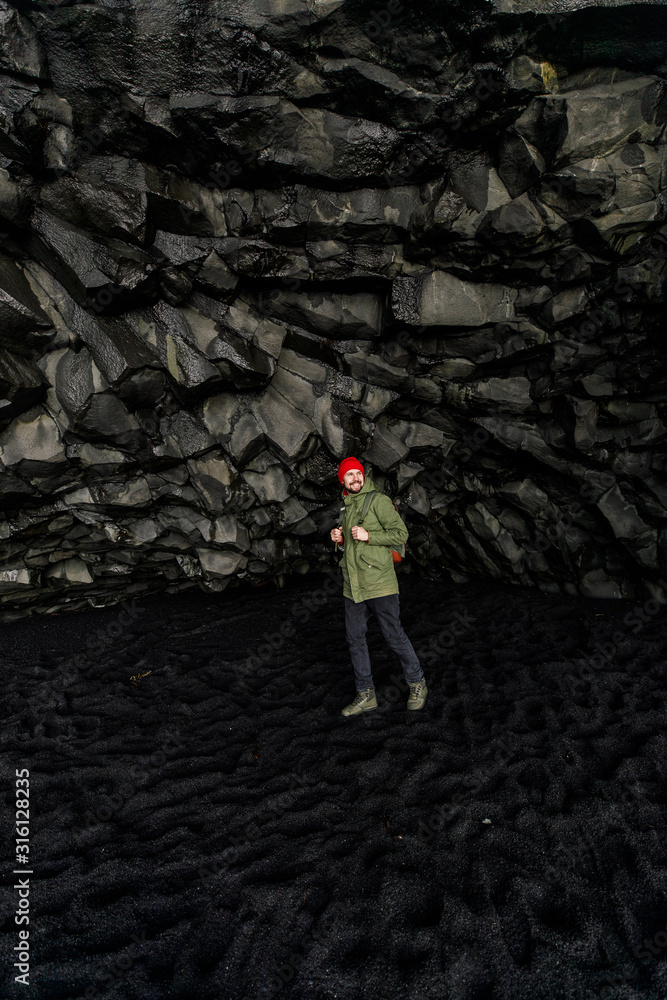 young male traveler in green clothes in a red hat and a green backpack in a black cave in Iceland