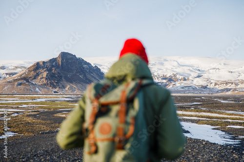young male traveler in green clothes in a red hat and a green backpack walks along the path against the background of mountains in Iceland