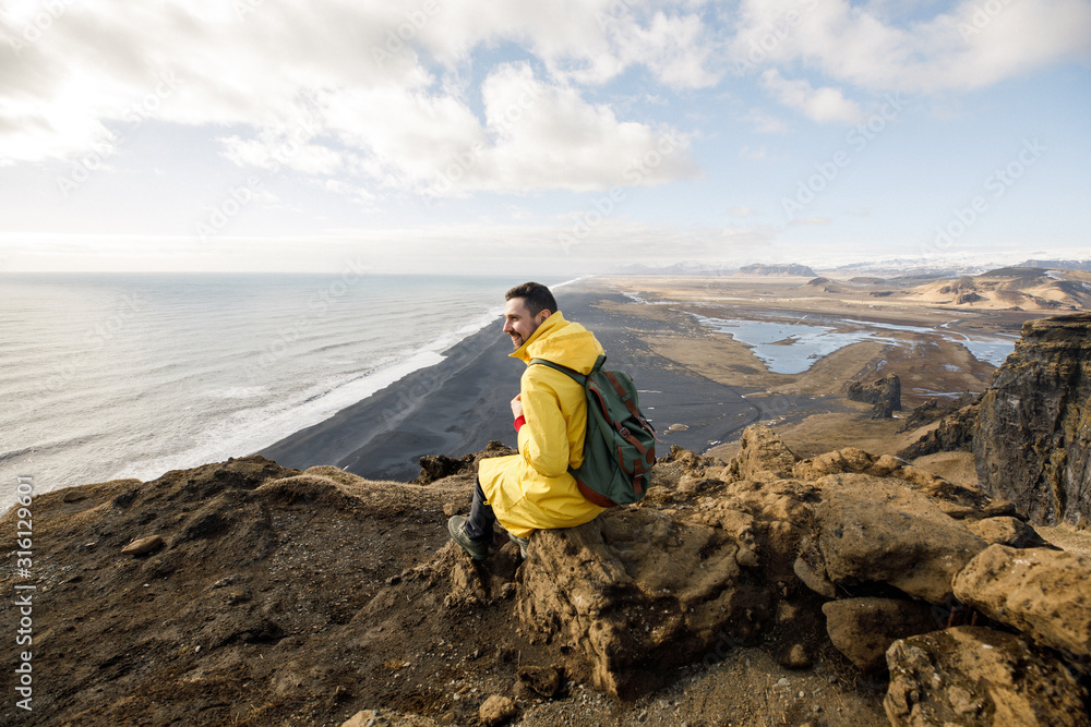 young male traveler in yellow clothes and a green backpack sitting near a cliff view of a black beach and the Atlantic Ocean in Iceland
