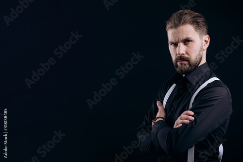 Half-turned portrait of masculine bearded man in black shirt and white suspenders isolated black background copyspace