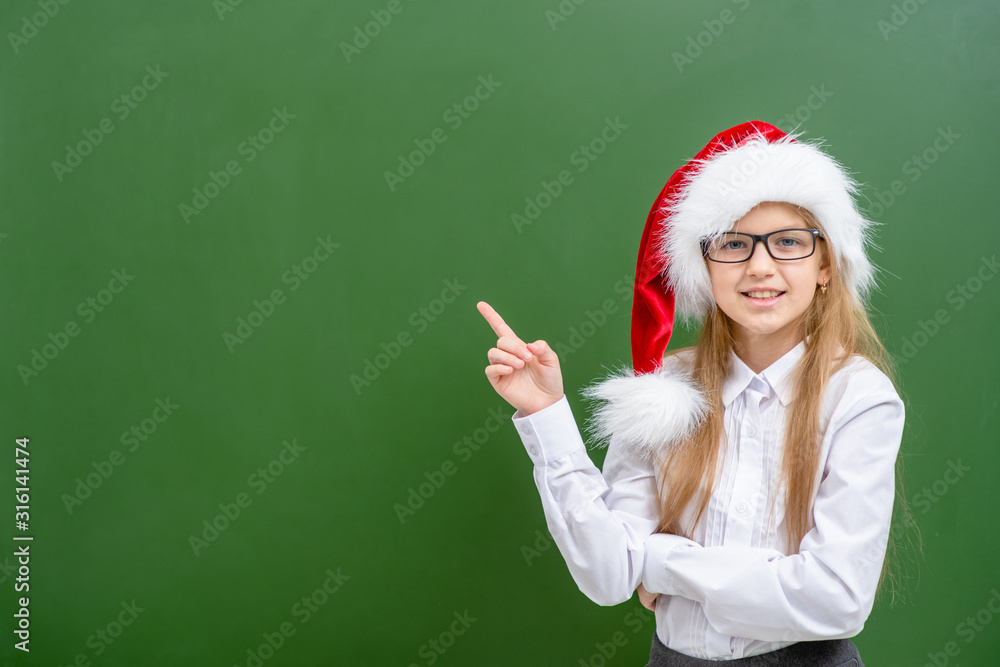 Smiling girl wearing a red santa hat and eyeglasses points with finger away on empty chalkboard. Space for text Smiling girl wearing a red santa hat and eyeglasses points with finger away on empty chalkboard. Space for text