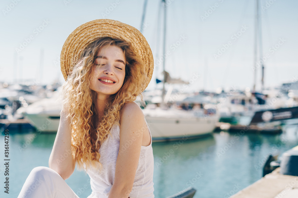 Girl covering her eyes in pleasure sits on a pier on the background of the ocean and ships