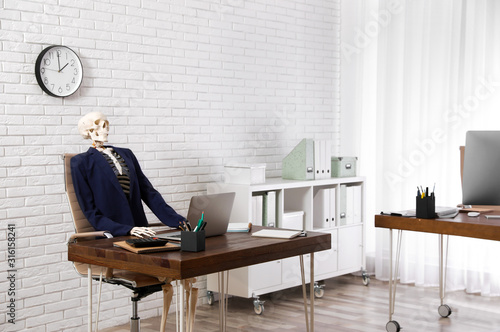 Human skeleton in suit using laptop at table in office