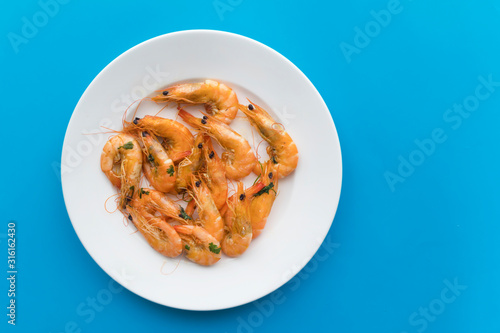 Cooked shripms, fried or boiled seafood, king tiger prawns with green herbs, spices on a white plate on a blue background. Top view. Healthy protein food, diet.