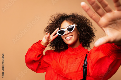  Portrait of beautiful young black woman taking selfie.Portrait of a pretty young afro american woman in retro style clothes smiling while standing and taking a selfie isolated over beige. photo