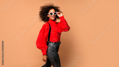 Horizontal fashion  image of excited black woman jumping with happy face expression on beige background. Wearing vintage red shirt