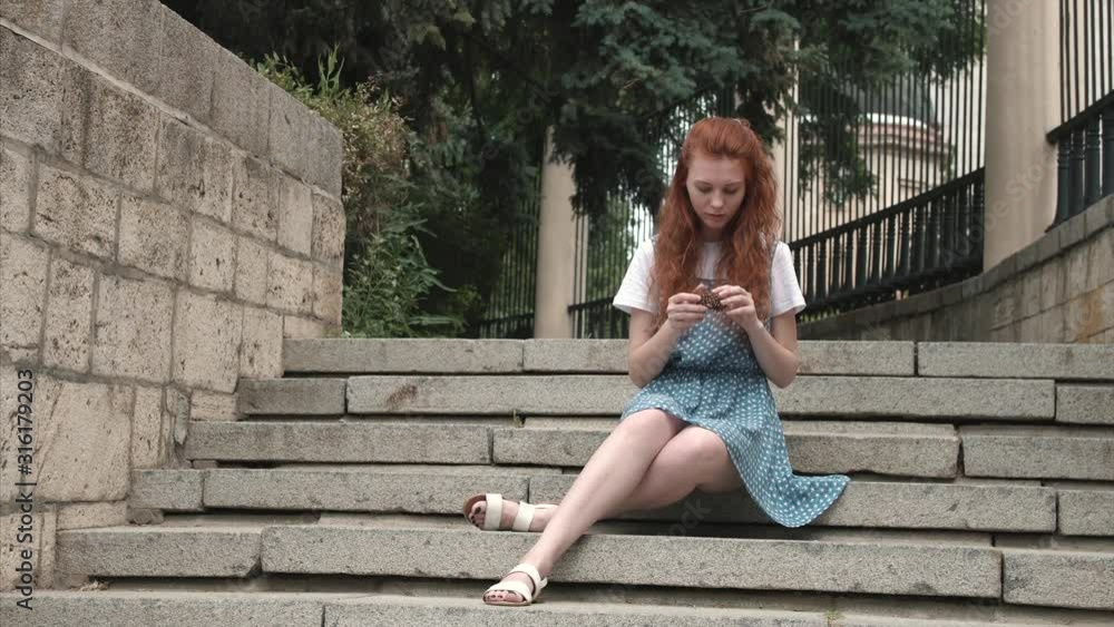 Cute redhead freckled teenager sitting on steps outdoor. Ginger girl touches fir cone. Beautiful red-haired woman in park. Cute redhead freckled teenager sitting on steps outdoor. Ginger girl touches fir cone. Beautiful red-haired woman in park.