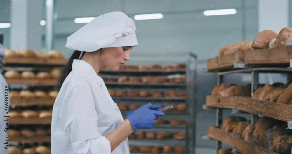 Beautiful lady baker take some notes in her tablet and photos of baked bread from shelves in a bakery industry Beautiful lady baker take some notes in her tablet and photos of baked bread from shelves in a bakery industry