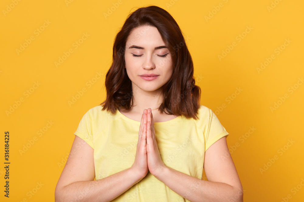Horizontal shot of young woman dresses casual clothes, posing isolated over yellow background in studio. People lifestyle concept. Female keeping hands folded in prayer, stands with closed eyes. Horizontal shot of young woman dresses casual clothes, posing isolated over yellow background in studio. People lifestyle concept. Female keeping hands folded in prayer, stands with closed eyes.