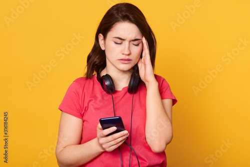 фотографія Close up portrait of woman with cellphone isolated over yellow studio background, attarctive female feels headache, young beautiful lady wears casual outfits, keeps eyes closed, touches temple