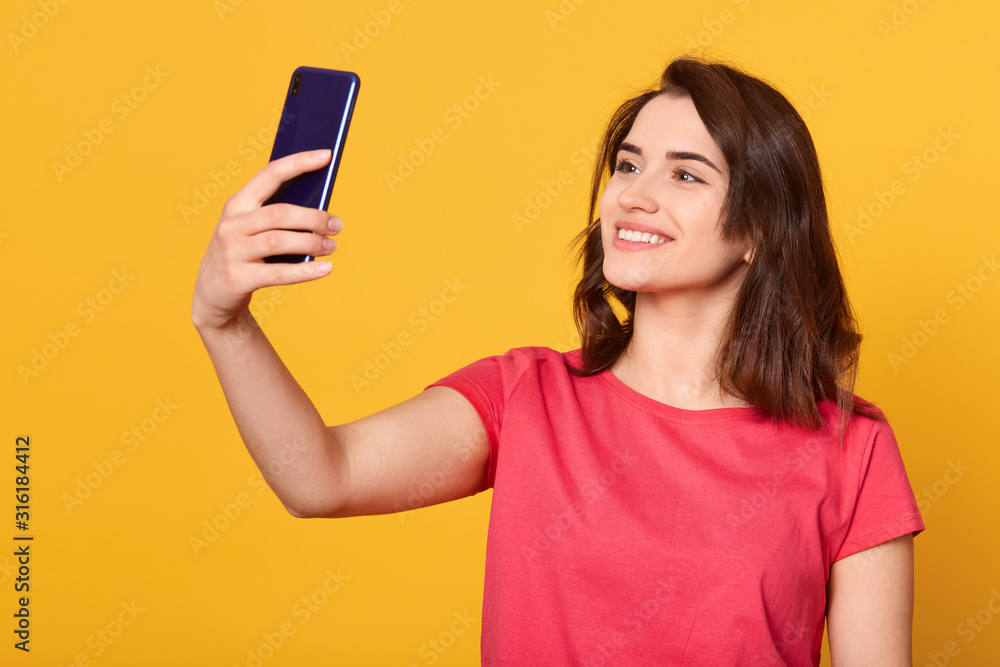 Horizontal shot of pretty girl taking self portrait with her smart phone, adorable lady looking devie's at camera with charming smile, attractive model posing isolated over yellow studio background. Horizontal shot of pretty girl taking self portrait with her smart phone, adorable lady looking devie's at camera with charming smile, attractive model posing isolated over yellow studio background.
