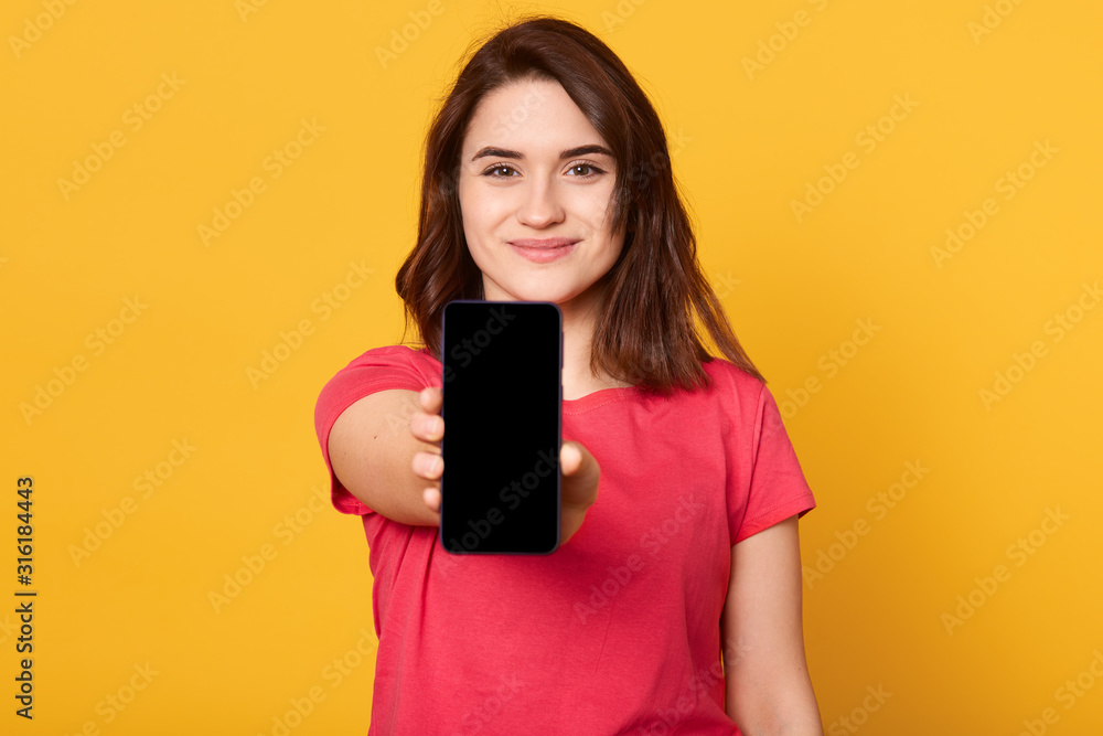 Close up portrait of young woman showing display of mobile cell phone with black blank screen and looks smiling directly at camera isolated over yellow background. Copy space for advertisment. Close up portrait of young woman showing display of mobile cell phone with black blank screen and looks smiling directly at camera isolated over yellow background. Copy space for advertisment.