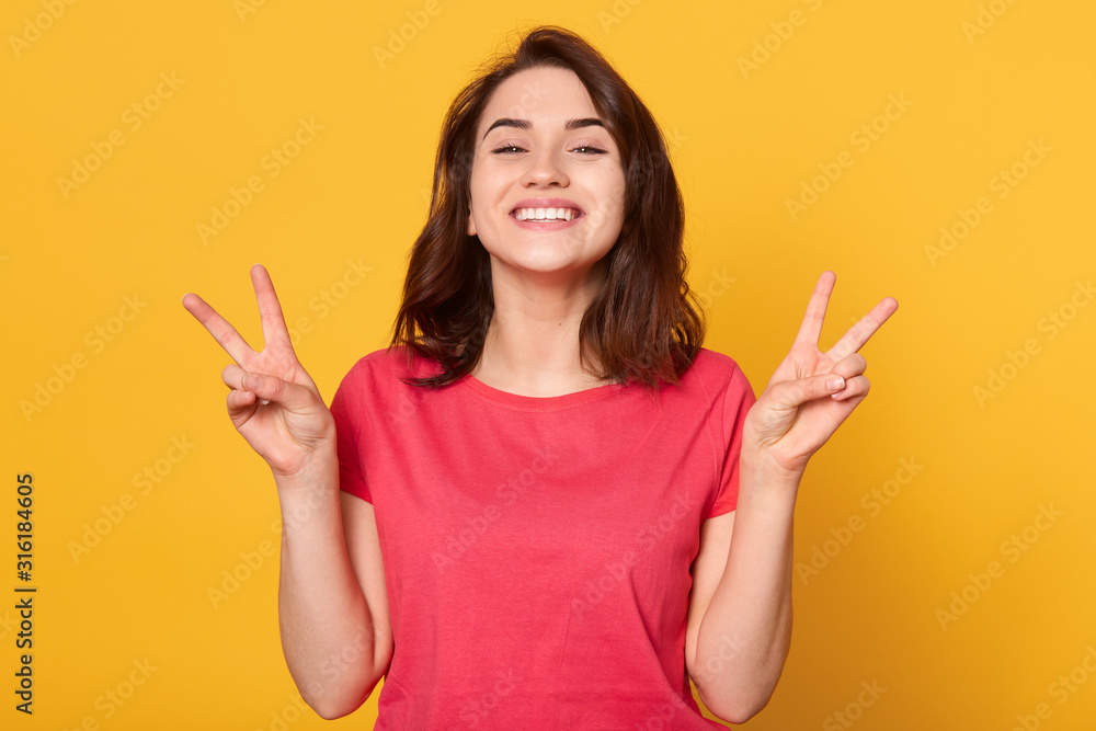 Indoor shot of young woman with dark hair posing in studio over yellow background, dresses bright clothes, showing victory sign, looks happy,looking directly at camera.People emotions concept. Indoor shot of young woman with dark hair posing in studio over yellow background, dresses bright clothes, showing victory sign, looks happy,looking directly at camera.People emotions concept.